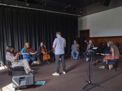 Violinists Meredith Bates and Josh Zubot, cellist Peggy Lee, violinist Wesley Hardisty, violinist Trent Freeman, and vocalist Pura Fé sit in a circle in the Grand Luxe Hall. Jesse Zubot stands in the centre, wearing a grey pocket T-shirt, baseball cap, and dark-washed jeans. He faces the musicians, with his back to the audience.