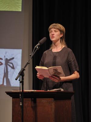 Klara du Plessis stands behind a lectern in the Grand Luxe Hall while holding an open copy of her book Ekke. She wears a black linen dress, and is speaking into a microphone on a stand while staring out at the crowd.