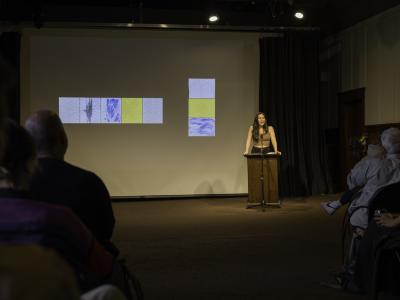 Deanna Fong stands behind a lectern in the Grand Luxe Hall, leaning on its surface with her elbows locked. Behind her is a projected image by Christian Vistan that is composed of eight water-based abstract paintings in yellow, white, and blue.