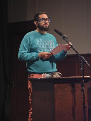 Khashayar Mohammadi stands behind a lectern and reads into a microphone that rests on a stand. They hold a stack of papers in their hands, and are wearing oversized glasses, a blue sweatshirt, and orange pants. A blue disposable mask, a leather wallet, and Apple headphones rest on the surface of the lectern.