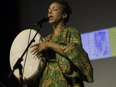 Tawhida Tanya Evanson vocalizes into a microphone in the Grand Luxe Hall while holding a drum. She wears a green and gold patterned dress with billowing sleeves.