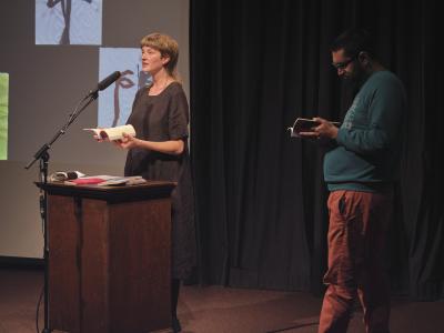 Klara du Plessis stands behind a lectern in the Grand Luxe Hall. She speaks into a microphone positioned on a stand while holding a book open with both hands. Klara gazes towards the audience, and wears a black linen dress. To her left, Khashayar Mohammadi gazes downwards towards the book they’re holding. They wear glasses, a blue sweatshirt, and orange pants.