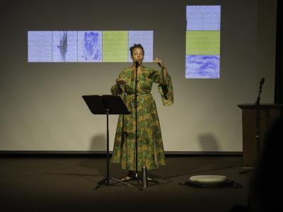 Tawhida Tanya Evanson gestures energetically while performing a poem. She wears a gold and green patterned dress and is barefoot in the Grand Luxe Hall. A music stand, microphone, silver thermos, and drum are positioned around her. A projected image of abstract paintings by Christian Vistan can be seen in the background.
