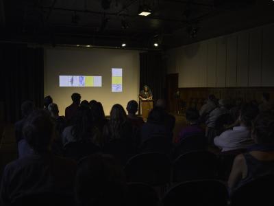 Junie Désil stands behind a lectern in the Grand Luxe Hall. Eight yellow, white, and blue abstract paintings by Christian Vistan are projected in the background. Rows of seated audience members can be seen from behind.