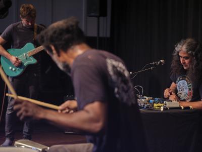 Marshall Trammell plays drums with his back to the camera. In the background, John Dieterich plays a turquoise blue electric guitar, and Raven Chacon plays synthesizer while seated at a table. 