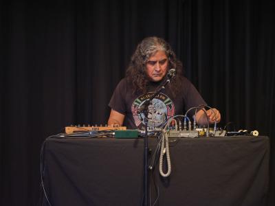 Raven Chacon sits at a table where various synthesizers, microphones, and effects pedals are arranged. He wears a black graphic T-shirt, and his hand hovers while adjusting one of his electronic instruments. 