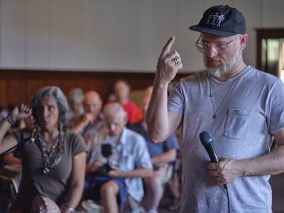 Jesse Zubot holds a microphone in his left hand, and points upwards with his right index finger. He wears a black baseball hat decorated with white embroidered people, glasses, and a silver pendant over a grey pocket t-shirt. In the background, Pura Fé vocalizes into a microphone and mirrors Jesse’s gesture with her right hand raised in a fist. The audience in the Grand Luxe Hall can be seen behind them.