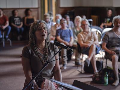 Pura Fé sings into a microphone with her eyes closed in the Grand Luxe Hall. She sits with her back to the audience, and wears colourful necklaces layered over a brown t-shirt.