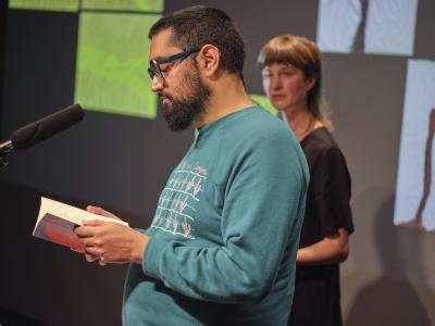 Khashayar Mohammadi wears a blue sweatshirt and blue oversized glasses. They gaze down towards a book held open in both of their hands. Klara du Plessis stands behind them, wearing a black linen dress.