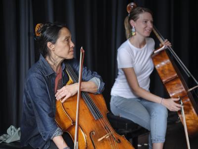 Cellists 
Peggy Lee and Marina Hasselberg sit with their instruments in the Grand Luxe Hall. Peggy wears a denim button-up and her hair is in a claw clip, and  Marina Hasselberg wears a white T-shirt, jeans, and geometric earrings. Her bow dangles between her fingers.