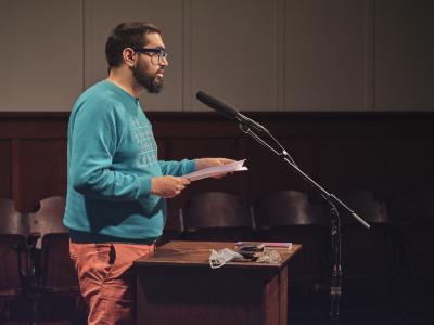 Khashayar Mohammadi is seen in profile standing behind a lectern in the Grand Luxe Hall. They hold loose sheets of printer paper and are reading into a microphone that is affixed to a stand. Khashayar wears oversized glasses, a blue sweatshirt, and orange pants. On the lectern, a copy of their book G is seen next to a wallet, blue disposable mask, and Apple headphones bundled in a knot.