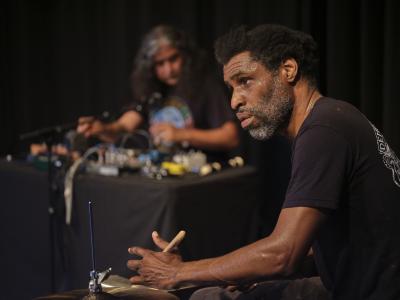 Marshall Trammell is seen in profile while playing drums. He wears a black t-shirt. Raven Chacon can be seen in the background at his table of synthesizers.