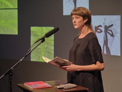Klara du Plessis stands behind a lectern in the Grand Luxe Hall. She wears a black linen dress, and holds an open copy of her book Ekke with both hands. On the lectern are two other books with red covers and a grey towel. She speaks into a microphone while gazing to the side. Abstract water-based paintings are projected in the background.