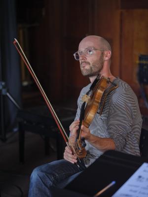 Trent Freeman gazes upwards while holding his violin in his left hand, and his bow in his right. He wears clear-framed glasses, a black-and-white striped shirt, and blue jeans.