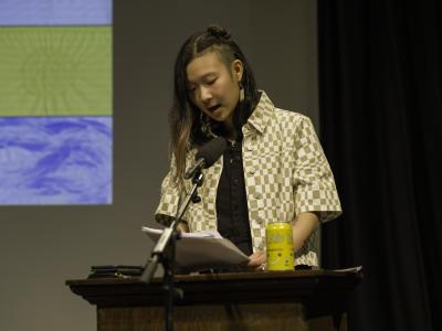 Isabella Wang gazes down at her notes at a lectern in the Grand Luxe Hall. She wears a cream and white checkered button up that is layered over a black shirt. A pineapple Bubly is positioned on the lectern next to her reading notes.