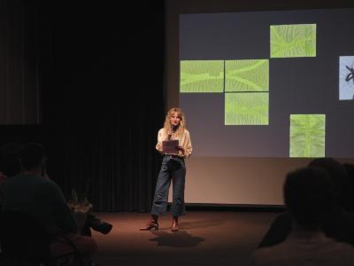 Lauren Lavery speaks into a cordless microphone while holding her notes in her left hand. She wears a white silk top, blue jeans, and brown boots. Behind her is a projected image by Christian Vistan of yellow water-based paintings arranged on a black background.