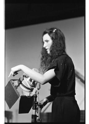 A medium-long side profile shot of a woman with long hair dressed in a jumpsuit. She points downward as her gaze looks off the image. Next to her is a stage light pointing down in the same direction of her gesturing hand.