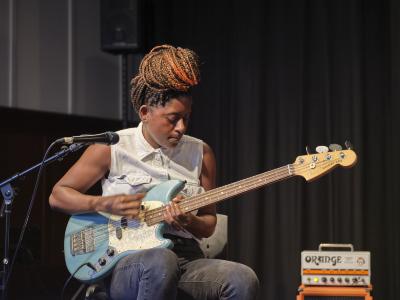 Farida Amadou plays a blue electric bass guitar. She gazes down at the strings, and is dressed in a white sleeveless button-up shirt, black jeans, striped socks, and black canvas sneakers. Her hair is worn in braids and tied in a top knot. A microphone on a stand is pointed towards her, and an Orange brand amplifier can be seen in the background.