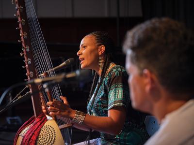 Depicted in profile, Sona Jobarteh holds her kora while speaking into a microphone. She wears her hair in braids, and is dressed in a black shirt with a blue pattern, blue jeans, beaded earrings, and a beaded cuff bracelet.