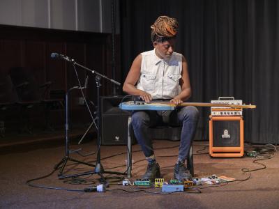 Farida Amadou sits in the Grand Luxe Hall with a blue electric bass guitar across her lap. She gazes down at the strings, and is dressed in a white sleeveless button-up shirt, black jeans, striped socks, and black canvas sneakers. Her hair is worn in braids and tied in a top knot. Various effects pedals and cables are arranged on the carpet around her feet, and a microphone on a stand is positioned to her right. A black speaker and an orange amplifier are positioned behind her.