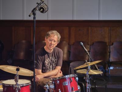 Dylan van der Schyff sits at a red drum kit in the Grand Luxe Hall. His arms are relaxed and crossed across his lap. Dylan wears clear-framed glasses, a black graphic t-shirt, and corduroy pants. 