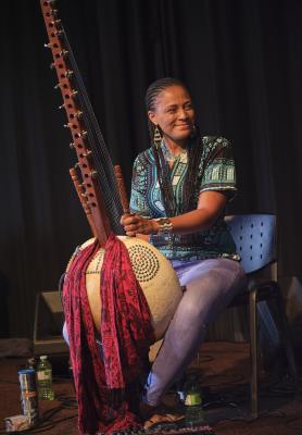 Smiling, Sona Jobarteh sits with her kora between her knees. She wears her hair in braids, and is dressed in a black shirt with a blue pattern, blue jeans, beaded earrings, and a beaded cuff bracelet. A red patterned scarf is wrapped around the neck of her instrument. A number of plastic water bottles, a thermos, and various cables are arranged on the carpet around Sona’s chair.