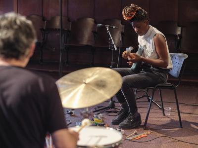 Farida Amadou is seated on a black armless chair while playing a blue electric bass guitar. She wears a white sleeveless button-up shirt, black jeans, striped socks, and black platform sneakers. Various effects pedals and cables are arranged on the carpet around her chair. In the foreground, Dylan van der Schyff is seen out of focus while playing drums with his back to the camera.