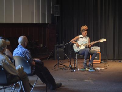 Farida Amadou plays a blue electric bass guitar in the Grand Luxe Hall. Her hair is worn in braids and tied in a top knot and she is dressed in a white sleeveless button-up shirt, black jeans, striped socks, and black canvas sneakers. A number of cables and effects pedals are arranged around her feet. Audience members seated in the first two rows can be seen from behind.