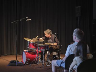Dylan van der Schyff plays a red drum kit. He wears clear-framed glasses, a black graphic T-shirt and dark pants. Audience members seated in the first two rows can be seen from behind.