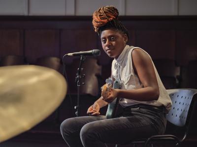 Farida Amadou plays a blue electric bass guitar while seated. Her hair is worn in braids and tied in a top knot and she is dressed in a white sleeveless button-up shirt and black jeans.