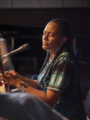 Sona Jobarteh plays kora with her eyes closed. She wears her hair in braids, and is dressed in a black shirt with a blue pattern, beaded earrings, and a beaded cuff bracelet. A microphone is pointed towards her.