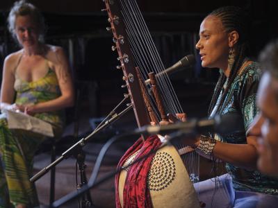 Depicted in profile, Sona Jobarteh holds her kora while speaking into a microphone. She wears her hair in braids, and is dressed in a black shirt with a blue pattern, blue jeans, beaded earrings, and a beaded cuff bracelet.