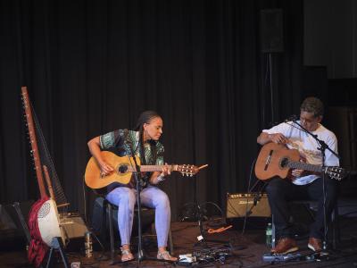 Sona Jobarteh and Eric Appapoulay play acoustic guitar while seated in the Grand Luxe Hall. Various guitar pedals are arranged on the carpet, and Sona’s kora is positioned in a stand to her right.