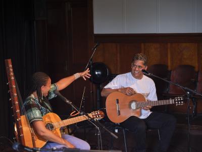 Sona Jobarteh and Eric Appapoulay sit with acoustic guitars in the Grand Luxe Hall. Sona points towards Eric, who smiles back in her direction.