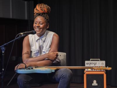 Farida Amadou smiles into a microphone while seated in the Grand Luxe Hall. Her blue electric bass guitar lies across her lap, and she leans on the instrument’s surface with her arms crossed. Her hair is worn in braids and tied in a top knot and she is dressed in a white sleeveless button-up shirt and black jeans. An Orange brand amplifier can be seen in the background.