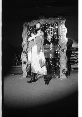 A performer basks in a spotlight wearing a thick beard, a dress of many colours and pointy black boots. They cross their arms, delicately resting their fingers at each shoulder. 
