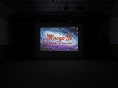 A still from a film projected in the Grand Luxe Hall. Mirage 0: Metanoia – Prelude is written in red stylized text and layered over a watercolour painting of a galaxy.