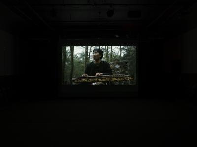 A still from a film projected in the Grand Luxe Hall that shows a man singing and playing a stringed instrument in a forest. The instrument is ornately carved, and laid horizontally across his lap. He wears a black shirt, and a black scarf wrapped around his head.