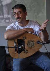 Aram Bajakian presents in the Grand Luxe Hall. He wears a white T-shirt, blue jeans, round glasses, and holds an oud.