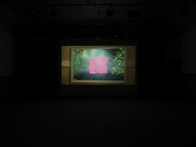 A still from a film projected in the Grand Luxe Hall. A framed picture of a lush oasis is overlaid with the title of the film in pink bubble letters. 