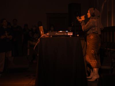 Rani Jambak stands behind a DJ table and sings into a microphone. She wears headphones over her head scarf, and is dressed in a sparkly top, long patterned skirt, and kitten heeled boots. The room is illuminated by a red light.