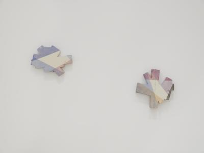 Two ceramic forms hang on a white wall. While they are different shapes, they’re similarly sized, and both resemble asterisks. Areas are painted in overlapping blue, pink, purple, and orange glaze using a masking technique. 
