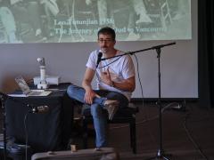 Aram Bajakian sits on a piano bench in the Grand Luxe Hall with his ankle crossed over his knee. He wears round glasses, a white t-shirt, blue jeans, and brown Birkenstock mules. He speaks into a microphone, and to his left is a table with a laptop and projector that is casting an archival photograph on the screen behind him.