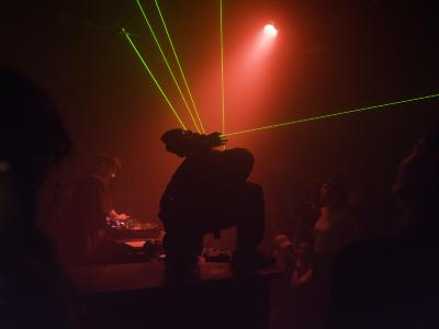 Ican Harem crouches on top of a DJ booth and shoots green lasers from his glove. Behind him, Kasimyn turns the dials on a CDJ. The room is illuminated in a red glow. 
