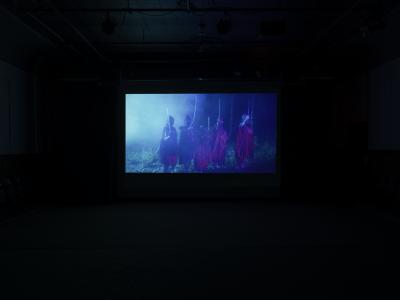 A still from a film projected in the Grand Luxe Hall. The image shows five young girls in a forest at night. They hold swords above their heads and are dressed in matching red traditional Indonesian clothing.