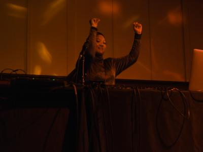 Rani Jambak stands behind a DJ table and dances with her arms up. She’s basked in a warm red light, and wears a head covering and sparkly long sleeved top.