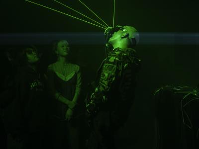 Ican Harm sings into a microphone as green lasers shoot from his glove. Two audience members are illuminated by the glow emitted from his hand.