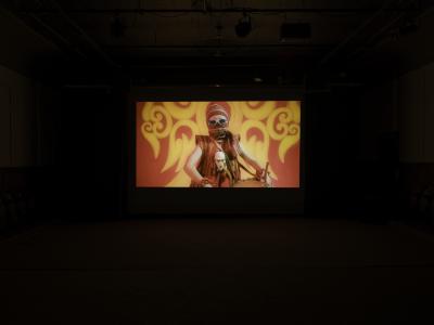 A still from a film projected in the Grand Luxe Hall. An image of a young girl dressed in sport sunglasses and red traditional Indonesian attire stands in front of a red patterned backdrop.