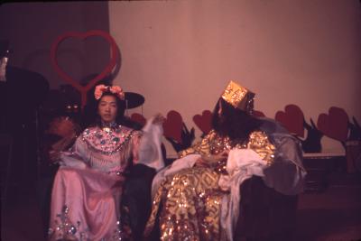 Paul Wong sits dressed in a pink satin gown decorated with blue and silver beading, another figure dressed in gold satin with sequinned detailing sits next to him. Paul's black hair is done up with pink roses which crown his demure, calm expressioned face. He's holding a white napkin in his left hand. Behind the figures heart shaped decorations held by Spirit Hands line the wall.