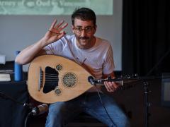 Aram Bajakian performs an oud demonstration. He is seated, and a microphone points to the body of the instrument. Aram wears a white graphic T-shirt, blue jeans, and round glasses.